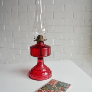 May include: A red glass oil lamp with a clear glass chimney. The lamp has a round base and a cylindrical body. A floral card rests on the table. The background is a white brick wall.