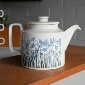 May include: A white ceramic teapot featuring a cylindrical body, a flat lid with a small knob, and a side handle. The exterior is decorated with a repeating pattern of blue line-drawn flowers and green foliage against a white background.