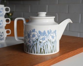 May include: A white ceramic teapot featuring a cylindrical body, a flat lid with a small knob, and a side handle. The exterior is decorated with a repeating pattern of blue line-drawn flowers and green foliage against a white background.