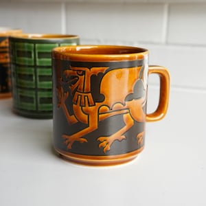 May include: A ceramic mug featuring a glossy amber-orange and matte black finish with a raised relief design of a mythical dragon creature. The mug has a cylindrical body, a sturdy handle, and a smooth rim. A second green mug is partially visible.