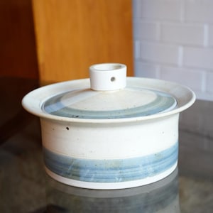 May include: A round ceramic lidded casserole dish featuring a speckled off-white glaze with horizontal bands of muted blue and grey. The lid includes a central cylindrical knob with a small circular opening, and the base has a wide rim.