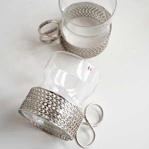 May include: Two clear glass cups with silver metal holders and handles. The holders have a textured, perforated design. The handles are a unique, looped shape. The cups are on a white surface.