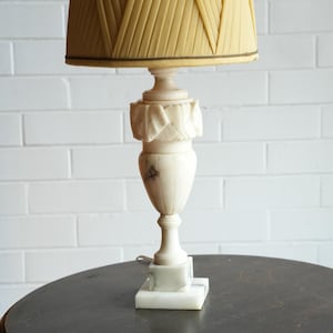 May include: A table lamp featuring a white marble base and body with carved leaf details, topped with a pleated, light yellow fabric lampshade. The lamp has a square base and a tapered, urn-shaped central column with visible grey veining.
