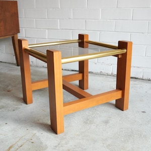 May include: A square side table featuring a light brown wooden frame with four thick vertical legs connected by horizontal crossbars. The top surface consists of a clear glass panel framed by four polished gold-tone metal cylindrical bars.