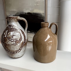 May include: Two ceramic jugs with handles. The left jug features a speckled off-white glaze with a dark brown swirling pattern. The right jug has a smooth, solid tan-brown glaze with a glossy finish. Both vessels have a rounded body shape.