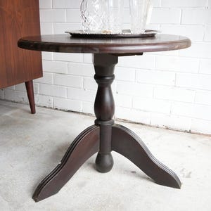May include: A round dark brown wooden pedestal table with a central turned column and three curved legs. On the tabletop sits a silver-coloured metal tray holding a clear glass decanter with a diamond-cut pattern and two matching drinking glasses.