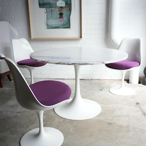 May include: A modern dining set with a round marble-topped table and three white tulip chairs with purple cushions. An abstract artwork hangs on the white brick wall. The table is approximately 100 cm in diameter.