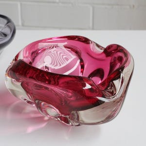 May include: A clear and ruby red glass ashtray with an organic shape. The ashtray has a deep red interior and a clear exterior, creating a layered effect. The design features rounded edges and a smooth surface.