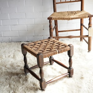 May include: A small, square wooden footstool with a woven, natural fibre seat. The dark brown legs are turned and angled. A tall wooden chair with a woven seat is in the background. The furniture sits on a white, shaggy rug.
