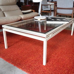 May include: A rectangular coffee table with a white frame and a clear glass top. The table features four straight legs and a tiered edge design. A white desk lamp and a black and white book are placed on the glass surface of the table.