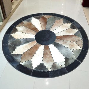 Può includere: Un tappeto circolare realizzato con pannelli di pelle bovina cuciti insieme in tonalità di nero, bianco, grigio e marrone. Il tappeto presenta un cerchio centrale nero circondato da un motivo a stella radiante di segmenti di pelle alternati.