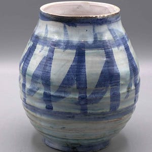 Op de afbeelding: Een keramische vaas met een rond lichaam en een brede opening, voorzien van een witte basisglazuur versierd met verticale, handgeschilderde blauwe penseelstreken en subtiele horizontale ribbels op het buitenoppervlak.