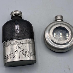 May include: A rectangular flask with a black leather upper section and a silver-toned metal base featuring an engraved floral pattern, alongside a circular silver-toned metal flask with a central circular glass window and a screw-top cap.