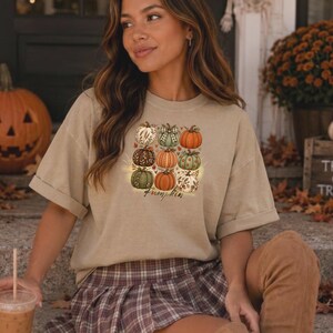 Puede incluir: Una camiseta de manga corta de color beige con un estampado gráfico de nueve calabazas estilizadas en varios colores y patrones, incluyendo naranja, verde y marrón, con la palabra "pumpkin" impresa en letra cursiva bajo el diseño.