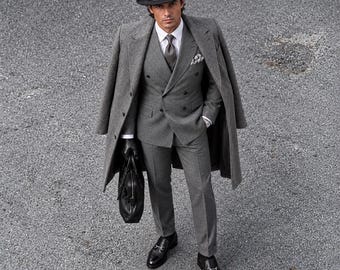 Può includere: Un abito in lana grigio antracite con giacca doppiopetto, pantaloni abbinati e camicia bianca con cravatta grigia. L'outfit include un lungo cappotto grigio, un cappello fedora nero, guanti in pelle nera, mocassini neri e una borsa nera.