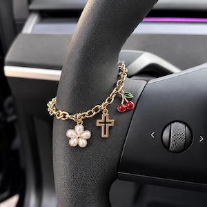 Puede incluir: Una pulsera de cadena dorada con tres dijes distintos: una flor blanca con una cuenta central tipo perla, una cruz dorada sencilla y un par de cerezas rojas con una hoja verde. La pulsera está colocada sobre un volante de color negro.
