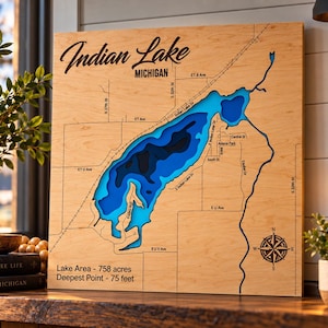 May include: A square wooden wall art piece featuring a laser-cut bathymetric map of Indian Lake, Michigan. The lake is depicted in layered shades of blue, surrounded by engraved street names and a compass rose. Text includes Lake Area - 758 acres and Deepest Point - 75 feet.