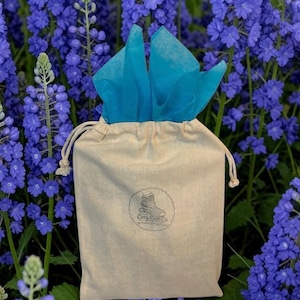 May include: A beige drawstring pouch made of fabric, featuring a circular logo with the text Cozy Cast Co. and a graphic of a cast. Bright turquoise tissue paper protrudes from the top opening of the bag, which is centered in the frame.