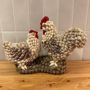May include: A decorative figurine featuring two chickens crafted from small white and brown seashells. The chickens have bright red shell combs and wattles, and they are mounted on a base covered in small, dark grey, rounded seashells.