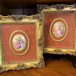 May include: Two rectangular gold-toned frames with ornate scrollwork detailing. Each frame contains a central oval plaque featuring a colorful illustration of a couple in period clothing set against a landscape, mounted on a dark orange fabric background.