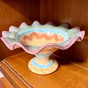May include: A ceramic pedestal bowl featuring a wide, ruffled rim painted in alternating bands of pastel pink, green, and blue. The interior displays a wavy orange pattern with yellow dots, while the base has a checkered yellow and cream design.