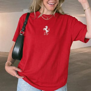 May include: A bright red short-sleeved t-shirt featuring a white embroidered prancing horse logo centered on the chest above the letters SF. The shirt is paired with a black shoulder bag and light blue denim jeans, creating a casual look.