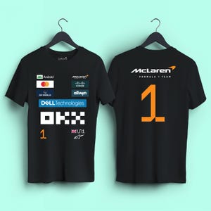 May include: Two black short-sleeve t-shirts featuring McLaren Formula 1 Team branding. The front displays various sponsor logos including Android, Mastercard, Dell Technologies, and OKX. The back features the McLaren logo and a large orange number 1.