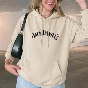 以下が含まれることがあります： 胸元にアーチ状のフォントでJack Daniel'sという黒い文字がプリントされたベージュのフード付きスウェットシャツ。前面にはカンガルーポケットがあり、長袖で、フードからは同色のベージュのドローストリングが垂れ下がっています。