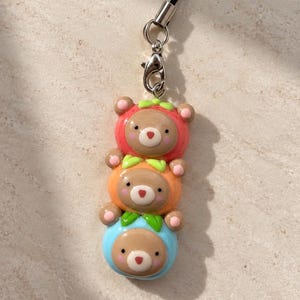 May include: A phone charm featuring three stacked brown bear figures, each wearing a different colored hood: red, orange, and blue. Each hood is accented with small green leaves at the top. The charm is attached to a silver metal lobster clasp and a black cord.