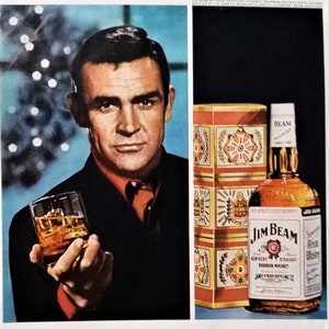May include: A man in a suit and red tie holds a glass of amber liquid. The text "The gift is distinctive. The season is merry. The man is Sean Connery. The Bourbon is JIM BEAM." is below the image. A bottle of Jim Beam bourbon whiskey is on the right side of the image.