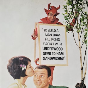 May include: A devil in a red costume holds a sign that reads "To build a man trap: Fill picnic basket with Underwood Deviled Ham Sandwiches". A woman is kissing a man on the cheek.