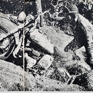May include: A black and white vintage photograph showing a motorcycle stuck in deep sand with a rider pushing it. The motorcycle has a prominent fuel tank and exposed engine components. Text at the bottom reads: throttle wide open, an earthbound cycle gets an assist from its rider. Jack Pine is not a race. Drivers must average 24mph between 44 check stations. Late or early a