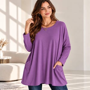 May include: A long-sleeved, loose-fitting purple tunic top featuring a round neckline and two front patch pockets. The garment has a relaxed, flared silhouette that extends past the waistline, crafted from a solid-colored, smooth fabric.