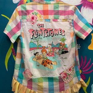 May include: A short-sleeved button-down shirt featuring a pastel plaid pattern and a large central graphic of The Flintstones characters. The back panel includes floral fabric patches and a pleated yellow hem. The shirt is displayed on a hanger.