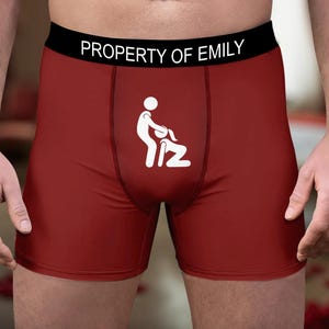 Puede incluir: Calzoncillos tipo bóxer de color rojo oscuro con una cinturilla negra con el texto blanco PROPERTY OF EMILY. La parte delantera de la prenda muestra una ilustración gráfica blanca de dos figuras de palo en una posición sexual.