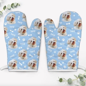May include: A pair of light blue oven mitts featuring a repeating pattern of a white dog, the name Daisy, and white paw prints and bone shapes. The mitts have a quilted texture and white trim along the bottom edges for a clean, finished look.