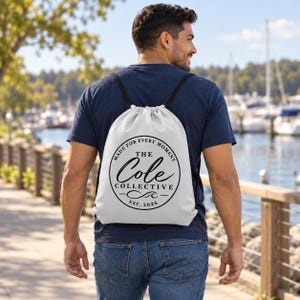 May include: A white drawstring backpack featuring a black circular logo with the text "MADE FOR EVERY MOMENT", "THE COLE COLLECTIVE", and "EST. 2026" in the centre. The bag has black cord straps and is worn over a dark blue t-shirt and blue jeans.