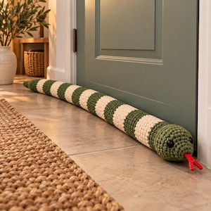May include: A long, cylindrical door draught excluder crocheted in a repeating pattern of olive green and cream-coloured stripes. The object is shaped like a snake with a rounded head, two small black bead eyes, and a bright red forked tongue.