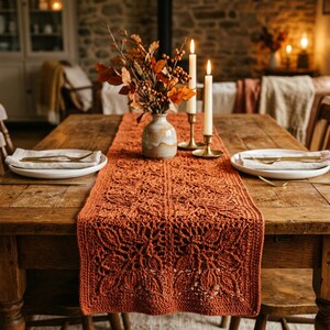 May include: A burnt orange crochet table runner featuring a repeating leaf pattern, laid flat on a wooden table. The runner is centered between two white plates with silverware and two lit taper candles in gold holders, with a vase of autumn foliage.