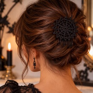 Peut inclure: Un peigne à cheveux noir orné d'un motif circulaire en crochet représentant une toile d'araignée. L'accessoire est fixé dans une coiffure relevée brune. Une boucle d'oreille pendante noire assortie avec une perle en forme de goutte est visible.