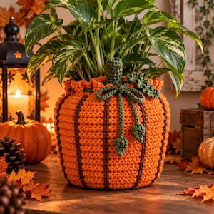 Puede incluir: Una funda de ganchillo para maceta de color naranja brillante con forma de calabaza, que presenta rayas verticales de color marrón oscuro, un tallo verde texturizado y un lazo decorativo de ganchillo verde con dos hojas pequeñas.