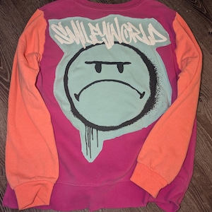 Peut inclure: Un sweat-shirt à manches longues avec un corps magenta et des manches orange vif. Le dos présente un grand graphique bleu clair d'un visage renfrogné avec des contours noirs et le texte "SMILEYWORLD" en police graffiti au-dessus.