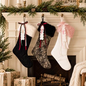 May include: Three Christmas stockings hanging in a row. The first is black lace with a white faux fur cuff and a tag reading Sophia. The second is black lace with a black faux fur cuff and a tag reading Elena. The third is white with a pink faux fur cuff and a tag reading Rosie.