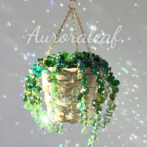 Könnte beinhalten: Ein hängendes Korb-Ornament mit herabhängenden Strängen aus facettierten Glasperlen in verschiedenen Nuancen von Limetten-, Smaragd- und Waldgrün. Der Korb ist aus hellem Material geflochten und an einer goldfarbenen Kette befestigt. Text: Auroraleaf.
