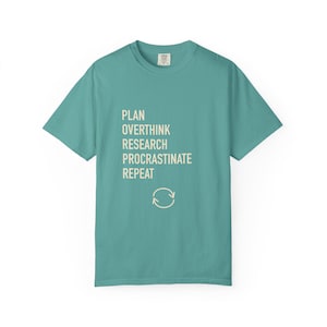 May include: A teal short-sleeved t-shirt featuring the cream-colored text PLAN, OVERTHINK, RESEARCH, PROCRASTINATE, REPEAT stacked vertically, followed by a circular arrow icon centered below the text on the front of the garment.