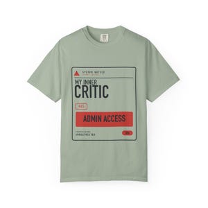 May include: A sage green short-sleeve t-shirt featuring a black and red graphic box with the text SYSTEM NOTICE, MY INNER CRITIC, HAS, ADMIN ACCESS, PERMISSIONS UNRESTRICTED, and a toggle switch labeled ON. The design mimics a computer system alert.