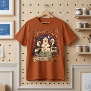 Può includere: Una t-shirt color ruggine con una stampa grafica che riporta il testo "CODE BOO CREW". L'illustrazione raffigura quattro anatre in costume di Halloween, tra cui una strega, un fantasma e un vampiro, attorno a un'anatra in un letto d'ospedale.