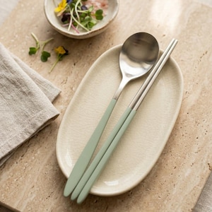 May include: A stainless steel spoon and a pair of chopsticks with matte sage green handles resting on an oval, cream-colored ceramic dish with a crackled glaze finish. The utensils feature a smooth, metallic finish on the top portions.