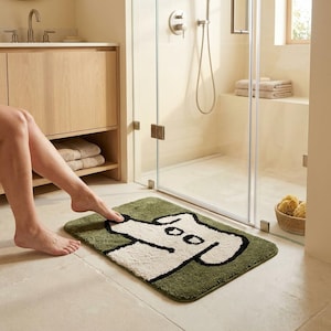 Può includere: Un tappetino da bagno rettangolare con un motivo a ciuffi raffigurante un cane bianco sporco con un bordo nero spesso su uno sfondo verde oliva a tinta unita. Il tappetino ha angoli arrotondati e una superficie morbida e testurizzata.
