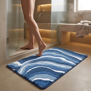 Op de afbeelding: Een rechthoekige badmat met een golvend, abstract patroon in tinten marineblauw, middenblauw, lichtblauw en wit. De mat heeft een dikke, getufte textuur met een donkerblauwe stoffen rand rondom de gehele omtrek van het kleed.